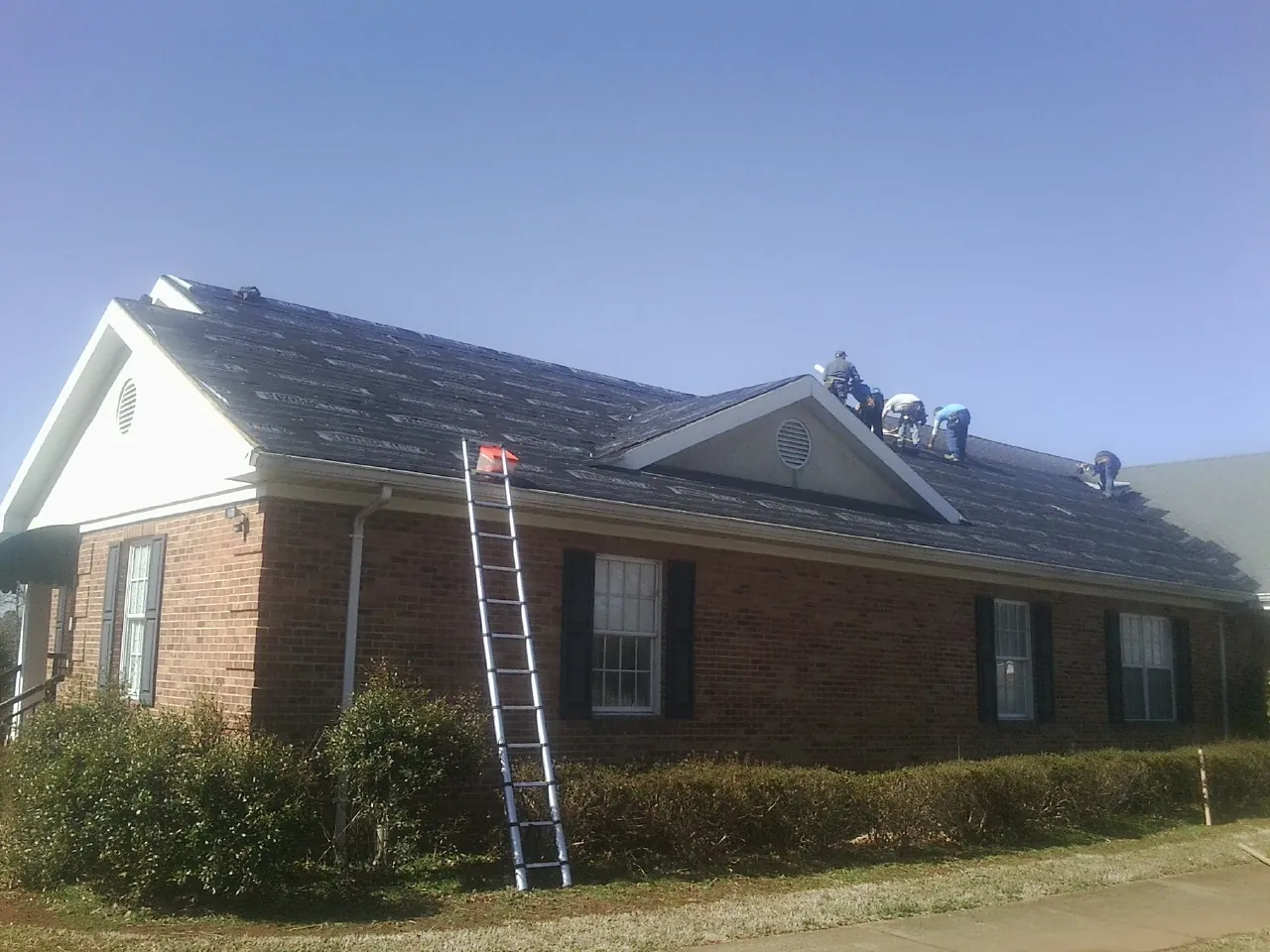 Crew tearing shingles off a brick ranch roof with a ladder set — Emergency Roof Repair work by our crew in Holtsville