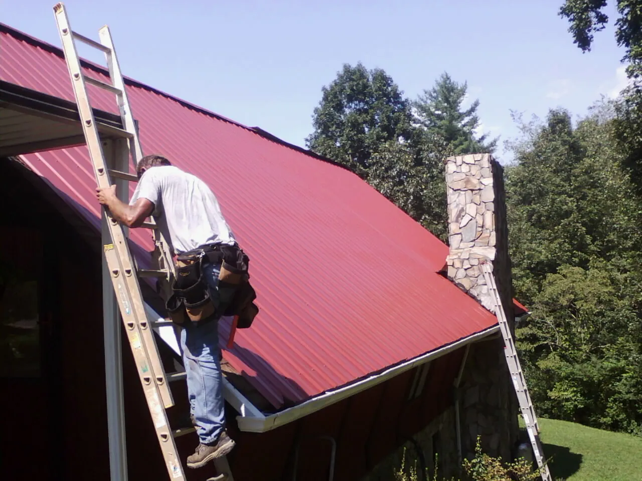Roofer on a ladder beside a red metal roof and stone chimney — Soffit & Fascia Repair by Roofing Brothers in Holtsville